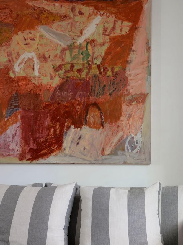 Close-up interior detail showing a large abstract red, terracotta, cream, and mauve artwork mounted on a pale wall above a grey upholstered sofa or daybed. Several oversized striped cushions in grey, white, and light taupe fill the foreground. The composition is quiet and contemporary, with the artwork providing a strong warm accent.