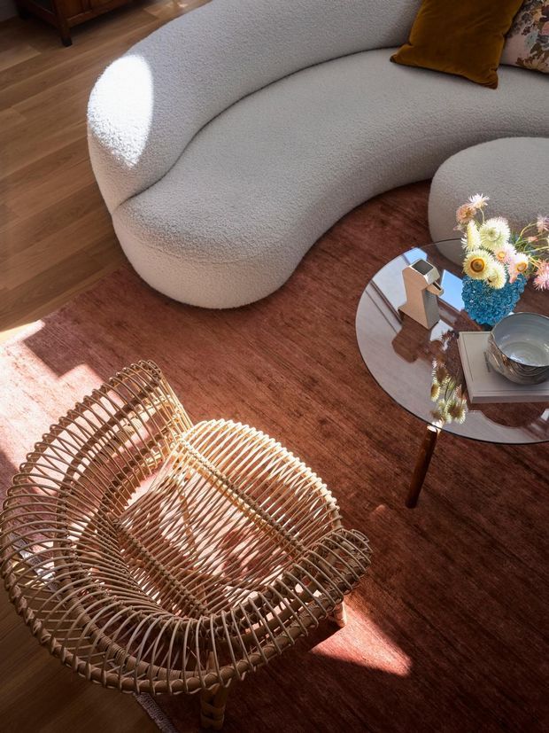 A warmly lit living room vignette featuring a sculptural curved off-white boucle sofa, a woven rattan lounge chair, a round reflective coffee table, and a rust-toned area rug over timber flooring. Decorative cushions, books, a metallic bowl, and a colourful floral arrangement add detail, while strong sunlight creates bright highlights and shadows.