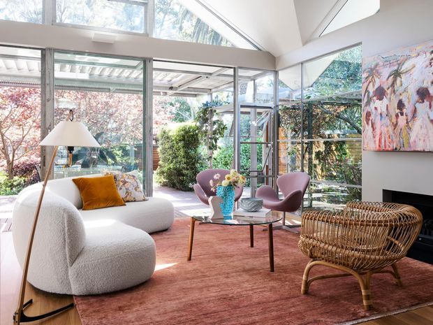 Bright, art-filled living room with expansive floor-to-ceiling glazing and clerestory windows opening visually to a lush garden. The room features a sculptural white boucle sofa, pink upholstered lounge chairs, a glass coffee table, a woven rattan armchair, a muted rose rug, and a contemporary fireplace with colourful artwork above it.