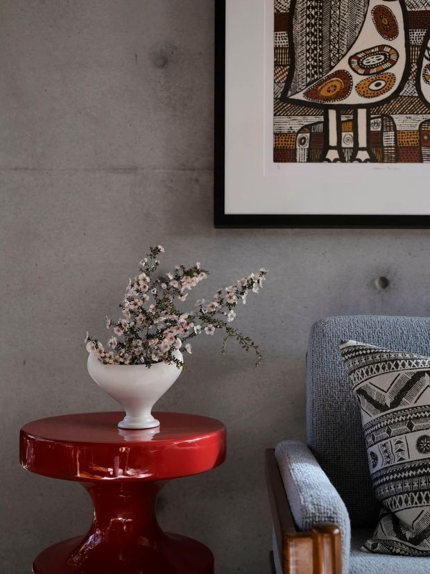 A close-up interior vignette featuring a glossy red pedestal side table topped with a white vase of delicate flowering branches. A textured blue-grey upholstered armchair with a patterned black-and-white cushion sits beside it. The backdrop is a raw grey concrete wall with a framed graphic artwork, creating a refined, eclectic and artistic atmosphere.