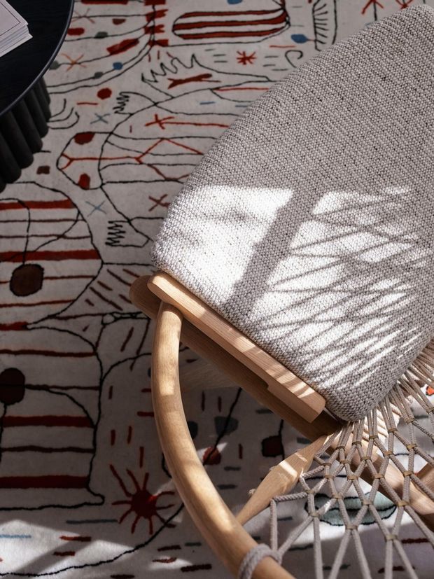 Close-up detail of a light wood-framed woven lounge chair with a grey-beige upholstered back and natural rope seat, positioned on an abstract patterned rug. Strong sunlight casts shadows across the chair, creating a warm, tactile interior vignette. A dark round side table is partially visible at the upper left.