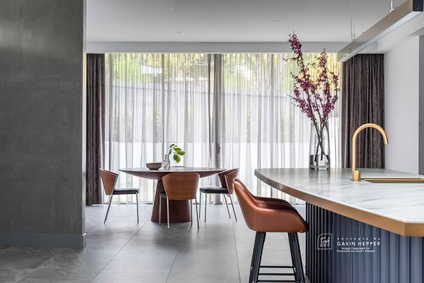 Contemporary open-plan dining and kitchen interior with a round timber dining table and sculptural chairs beside a large island. The space features pale large-format tiled flooring, dark textured wall surfaces, fluted dark island cabinetry, a light stone benchtop, brushed brass tapware, sheer full-height curtains, and abundant filtered daylight.