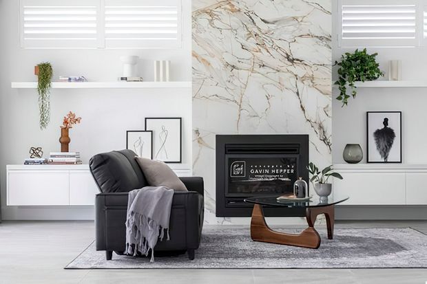 Bright contemporary living room featuring a black leather armchair with a draped throw, a sculptural wooden coffee table, low white built-in cabinetry, open shelving, wall art, and a central fireplace set into a dramatic white marble-look feature wall. White plantation shutters and a soft grey rug complete the refined neutral scheme.