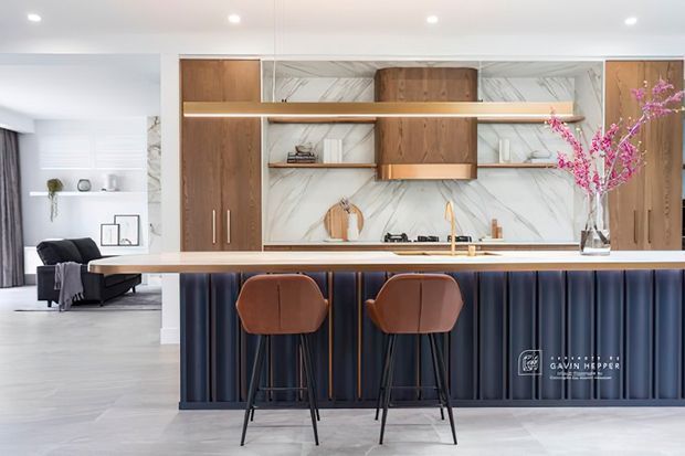 Bright contemporary open-plan kitchen with a large island and two bar stools. The kitchen features warm timber cabinetry, white marble-look wall cladding, integrated shelving, a dark fluted island base, and a slim illuminated shelf. A living area is visible beyond the kitchen, with minimalist furnishings and neutral finishes.