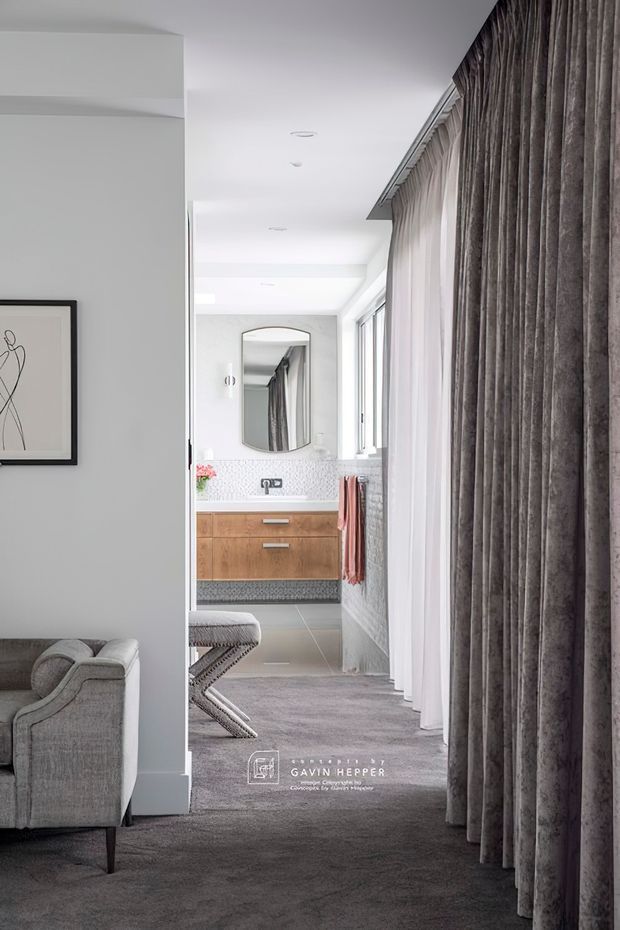 A refined interior view looking from a carpeted living area toward a bright bathroom. The foreground features a grey upholstered sofa, framed line artwork, and full-height textured grey curtains. The bathroom has a timber floating vanity with brushed metal handles, patterned mosaic wall tiles, a round-edged mirror, white walls, and a towel rail. The overall scheme is soft grey, white, and warm timber with a contemporary, polished feel.