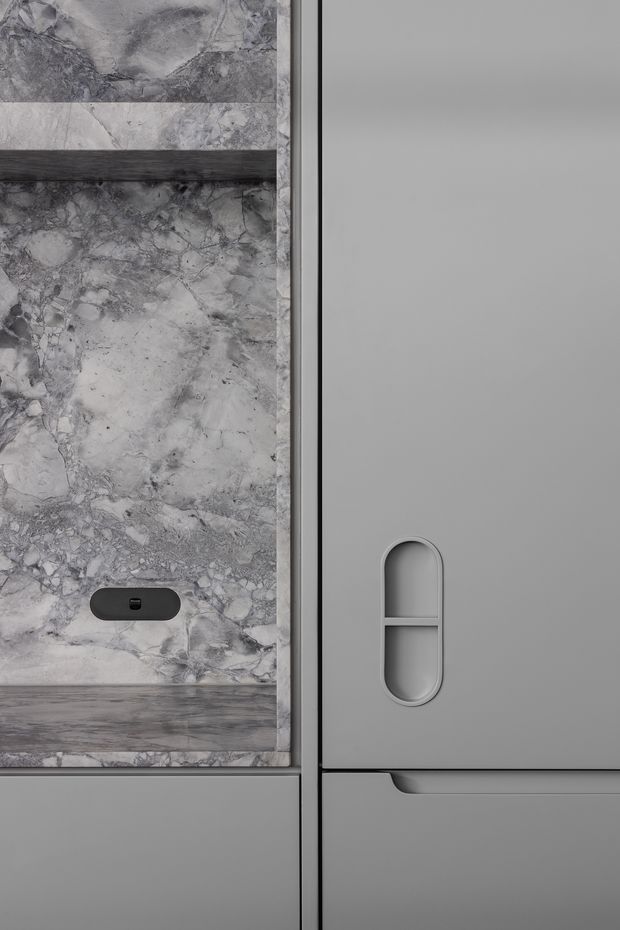 Close-up detail of a refined grey-toned built-in joinery installation. The left side features heavily veined grey natural stone panels with a dark horizontal timber or wood-look shelf and a small black recessed fitting. The right side shows smooth matte grey cabinetry with a sculptural recessed vertical handle and an integrated drawer pull. The composition is minimalist and monochromatic, focusing on material contrast and precise detailing.