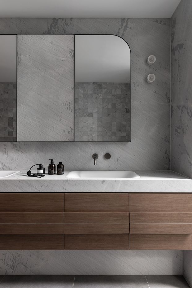Contemporary bathroom vanity with a long white integrated basin, dark timber drawer cabinetry, grey stone-look wall and floor tiles, black fixtures, and mirrored wall cabinets including a rounded corner mirror.