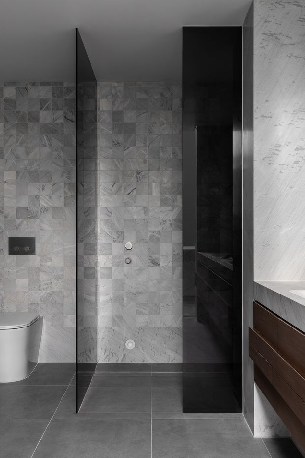 A contemporary bathroom featuring a frameless glass shower enclosure, grey patterned stone wall tiles, dark grey floor tiles, a wall-hung toilet, and a dark timber floating vanity with a pale stone benchtop. The palette is monochromatic grey and black with warm timber cabinetry accents.
