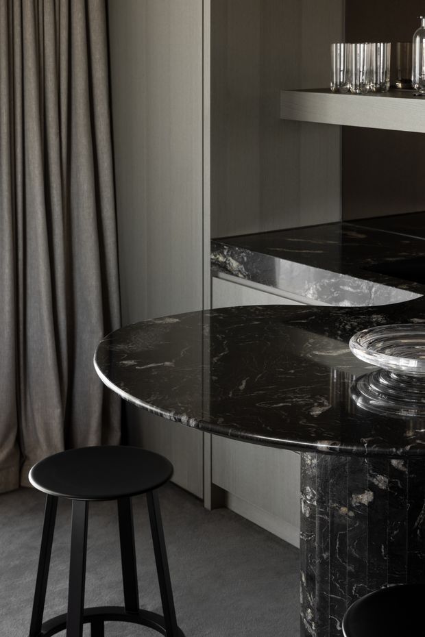 A moody, close-up interior view of a sophisticated kitchen or bar area. A rounded black marble peninsula with a heavily veined surface and matching waterfall support dominates the foreground, paired with a simple black bar stool. Dark cabinetry, a raised shelf displaying glassware, floor-to-ceiling grey curtains, and a grey floor create a refined monochromatic setting. A reflective vertical panel separates or mirrors the cabinetry and countertop.