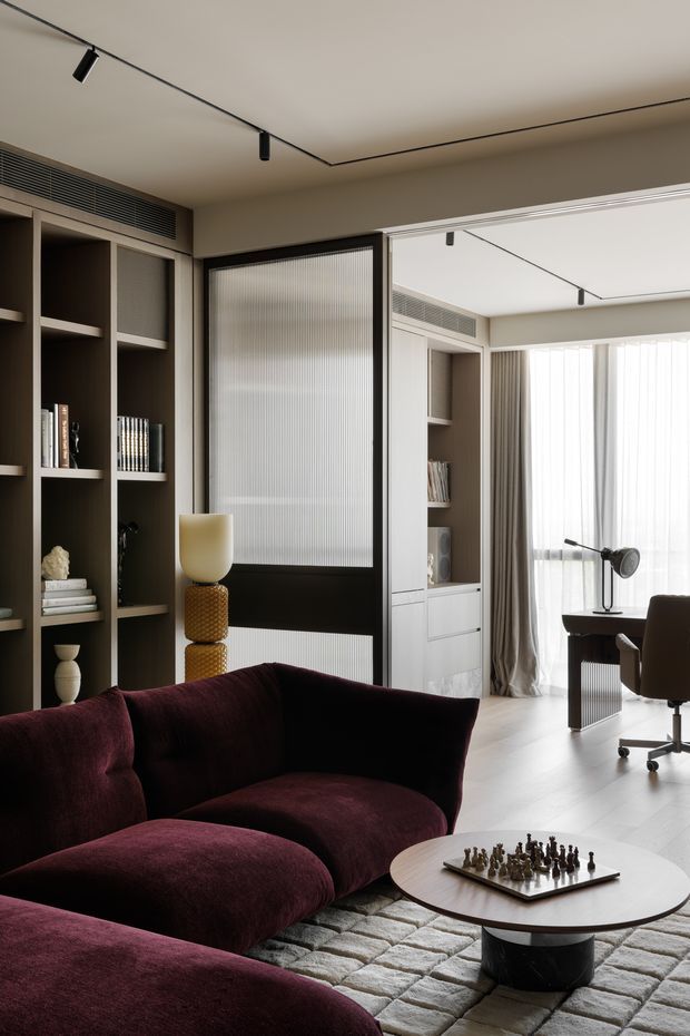 A refined contemporary living room with a deep burgundy sectional sofa in the foreground, a round coffee table with a chess set, built-in dark shelving, and a black-framed ribbed-glass sliding or pivot door. The room opens visually toward a bright workspace with a desk, task lamp, office chair, sheer curtains, and full-height glazing. Minimal black track lighting, neutral walls, pale flooring, and tailored drapery create a sophisticated, moody yet daylight-filled interior.