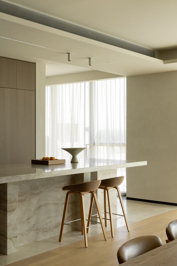 A refined, minimalist open-plan kitchen and dining area with a large pale stone island, integrated light-wood cabinetry, and timber bar stools. Soft daylight filters through full-height sheer curtains, while recessed linear air-conditioning vents and slim track lighting create a clean contemporary fit-out. A dining table and upholstered chairs are partially visible in the foreground.