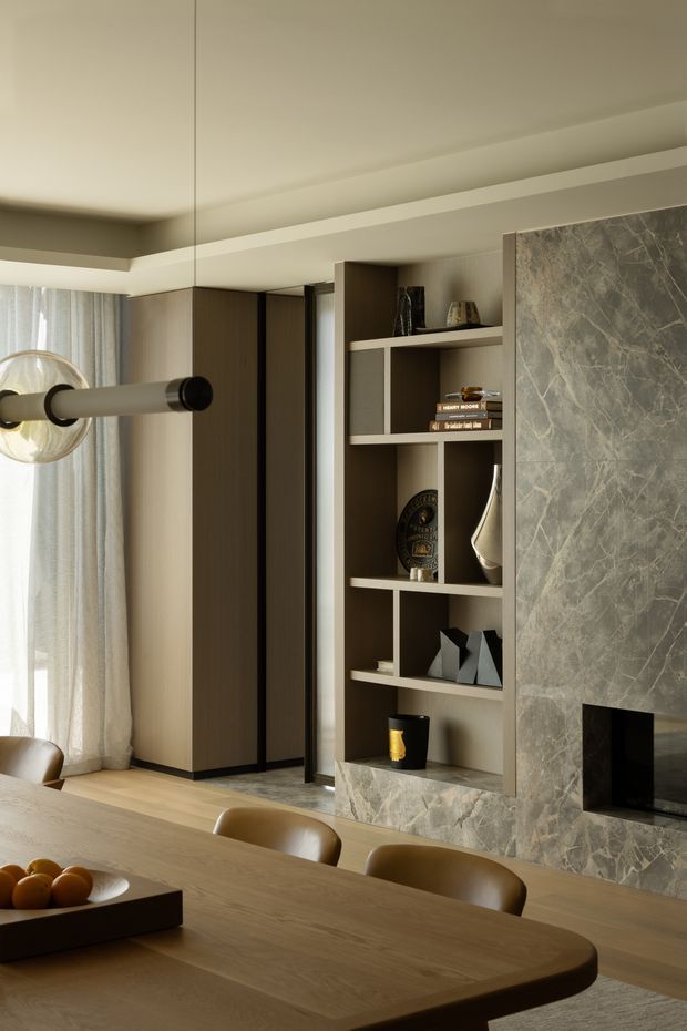 A warm, refined open-plan dining and living interior. A large timber dining table with curved edges and upholstered chairs occupies the foreground, while a built-in dark timber shelving unit displays decorative objects beside a substantial grey veined stone fireplace surround. Floor-to-ceiling curtains, pale timber flooring, concealed linear ceiling detailing, and a sculptural tubular pendant create a contemporary, softly layered space.