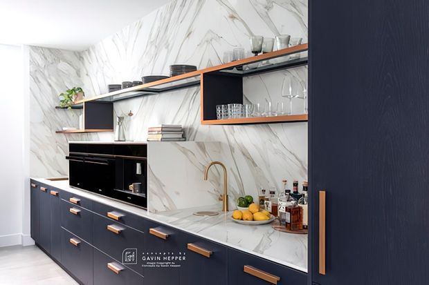 A compact contemporary kitchen with dark navy flat-panel cabinetry, warm timber or copper-toned pulls, open shelving, integrated black appliances, and a large white marble-look surface with grey veining wrapping the walls and benchtop. Glassware, cookware, and accessories are displayed on the open shelves and countertop.