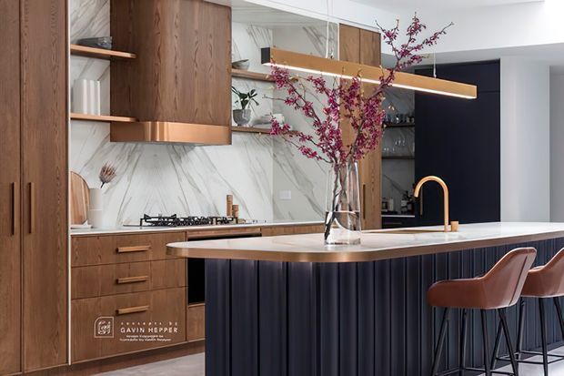 Contemporary kitchen featuring warm timber cabinetry, a white marble-look splashback, dark fluted island cabinetry, and a light stone benchtop. A copper-toned tap, integrated cooktop, open shelving, pendant lighting, and bar stools create a refined, high-contrast scheme.