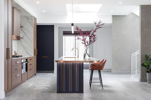 A bright, contemporary kitchen with a central island featuring a dark vertically fluted base and warm timber-toned benchtop. The kitchen includes full-height taupe cabinetry, integrated appliances, a marble-look splashback, pendant lighting, skylight, pale flooring, and a dark internal door. A small dining or sitting area and stair railing are visible in the background.