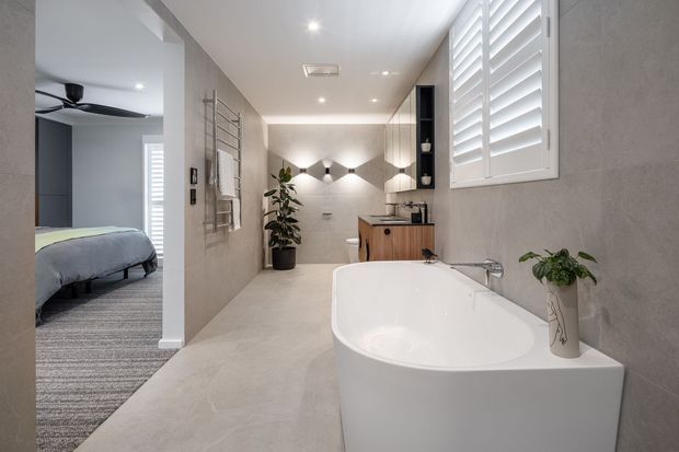 Contemporary bathroom featuring a freestanding white bathtub in the foreground, a walk-in shower at the rear, timber vanity cabinetry, pale stone-look wall and floor finishes, wall-mounted lighting, and white plantation shutters. An adjoining bedroom is visible through the open doorway.