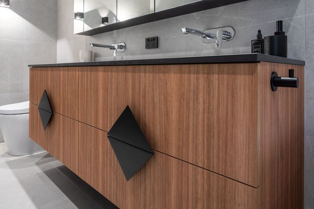 Close-up of a contemporary bathroom vanity with warm vertical-grain timber cabinetry, a dark stone benchtop, black geometric drawer pulls, and wall-mounted chrome tapware. A mirror, grey tiled walls, toilet, and grey tiled floor are visible.