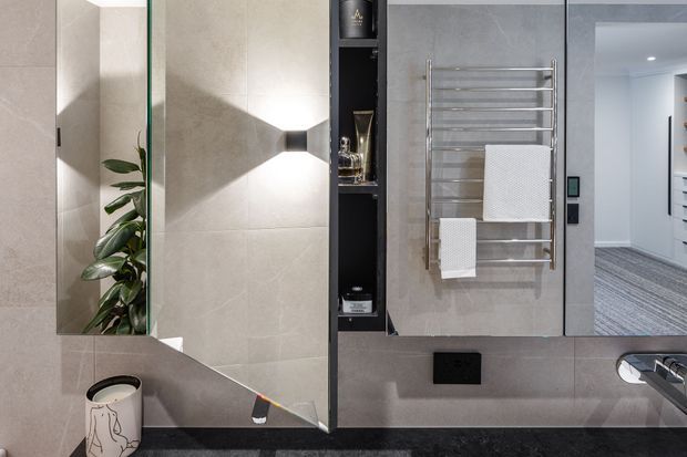 Contemporary bathroom vanity area with large pale stone-look wall tiles, a mirrored cabinet reflecting a leafy plant, open dark shelving, a wall-mounted chrome heated towel rail with white towels, and a dark stone benchtop with basin tap. A doorway reveals a carpeted adjacent room or dressing area.
