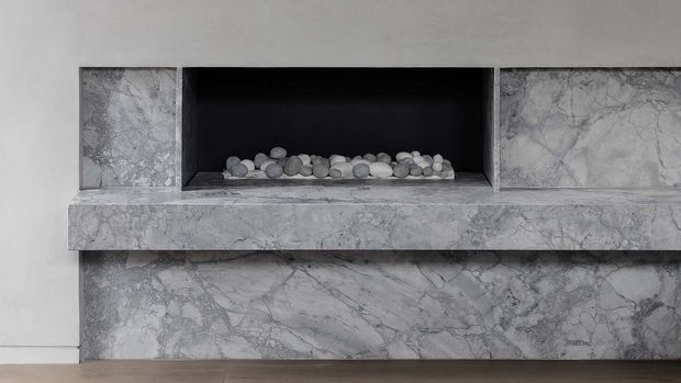 Close-up of a contemporary fireplace surround and hearth clad in heavily veined grey-white natural stone. The recessed firebox is finished in black and contains decorative rounded stones, with a simple white wall and timber floor visible around it.