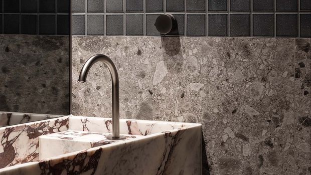 Close-up of a refined bathroom vanity featuring a sculptural marble basin with dramatic dark veining, a brushed metal gooseneck tap, a grey natural-stone wall finish, and contrasting dark square tiles above.