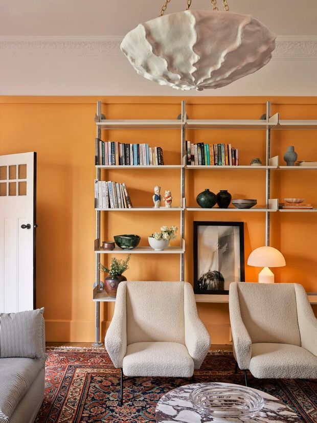 A richly styled living room with a vivid orange painted feature wall, open metal-framed shelving filled with books and decorative objects, two cream boucle lounge chairs, a partially visible sofa, layered Persian-style rugs, framed artwork, and a sculptural shell-shaped pendant light. The room combines warm colour, vintage furnishings, and eclectic decorative details.
