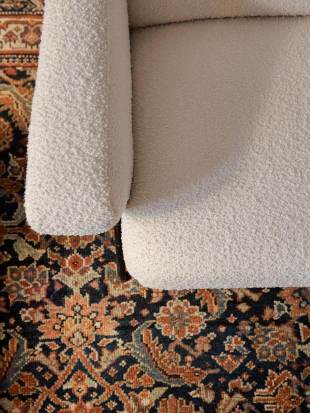 Close-up overhead view of a cream boucle-upholstered sofa or armchair with rounded edges, positioned on a richly patterned Persian-style rug in dark navy, rust, beige, and cream tones. The image focuses on upholstery texture, curved seating forms, and the contrast between the neutral furniture and ornate rug.