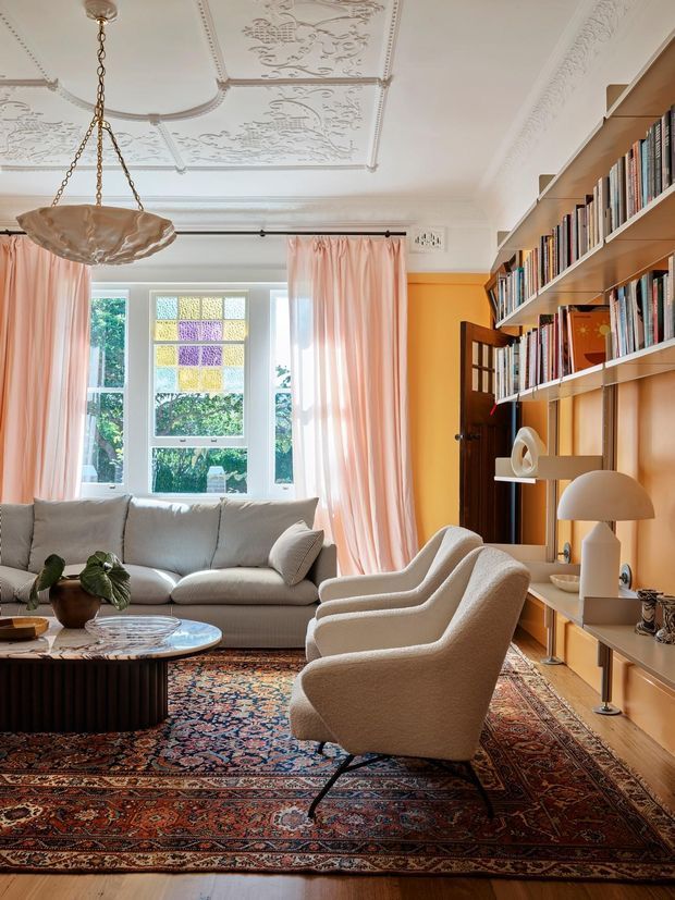 A warmly styled living room with an ornate decorative plaster ceiling, pendant light, tall stained-glass and clear-glazed windows dressed in blush-pink curtains, a cream sofa, sculptural cream lounge chairs, and extensive built-in timber shelving filled with books. A richly patterned Persian-style rug anchors the seating area around a low round coffee table, while a timber-framed glazed door opens at the rear.