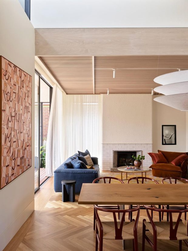 A bright, refined open-plan living and dining area with a warm timber palette. The room features a large solid timber dining table with sculptural timber chairs, a deep blue sofa, a rust-coloured lounge chair, herringbone timber flooring, a pale brick fireplace, full-height glazing with sheer curtains, and a timber-lined ceiling with integrated track lighting. Contemporary artwork and layered furnishings add character.