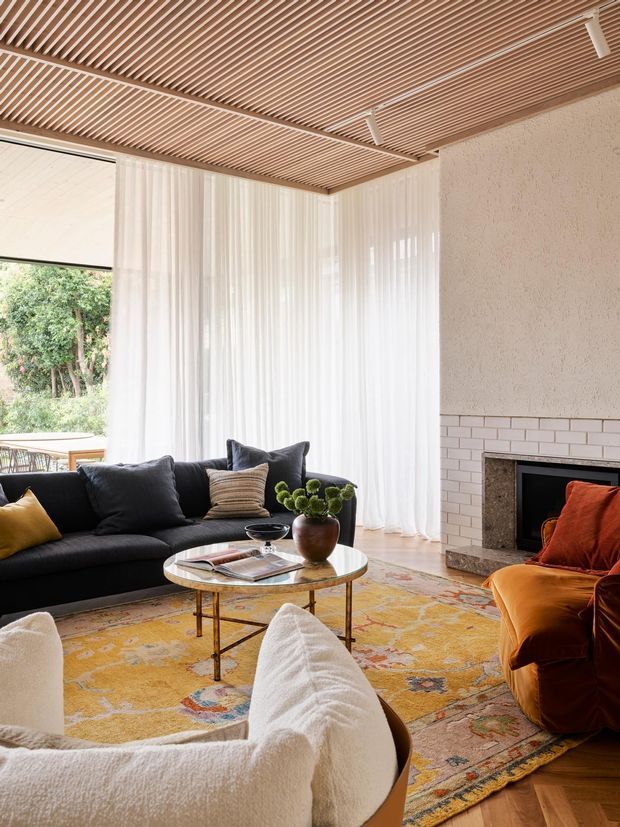 A warmly styled living room with a black sofa, sculptural cream lounge chairs, a rust-orange armchair, round glass coffee table, and a large patterned area rug. The room features sheer white curtains, a textured pale fireplace wall with white brick detailing, timber slatted ceiling, and abundant natural light from large glazing.