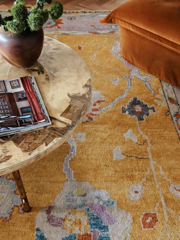 Close-up interior detail featuring a richly patterned mustard-gold area rug with abstract geometric motifs, a round marble-topped coffee table with a decorative veined surface, a dark brown vase holding green foliage, a magazine, and a burnt-orange upholstered ottoman or sofa edge. The composition has a warm, eclectic and layered decorative character.