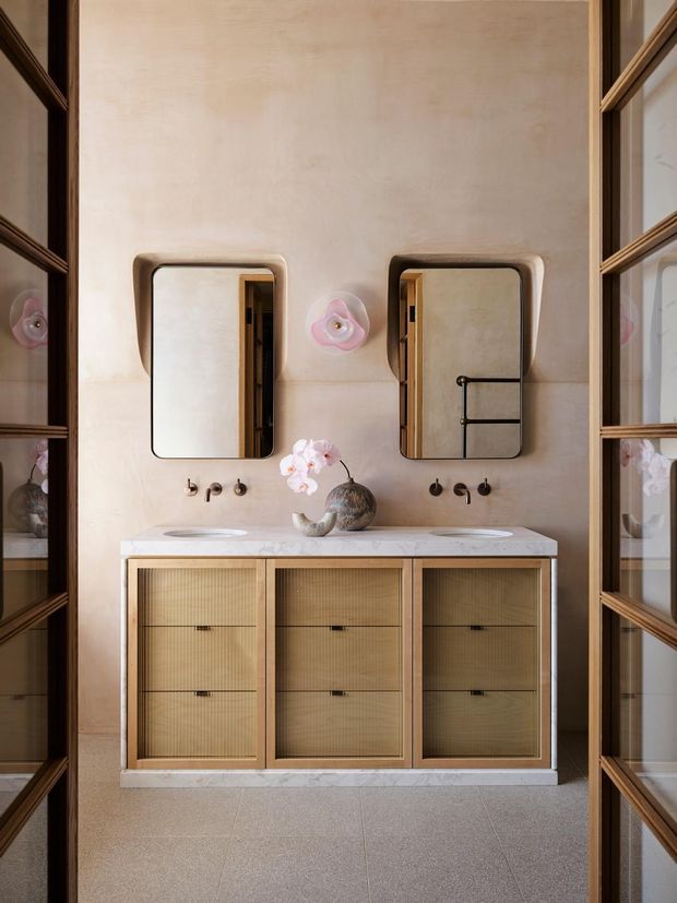 A refined double-vanity bathroom with warm plaster walls, a pale stone benchtop, ribbed timber cabinetry, bronze-toned fixtures, rounded rectangular mirrors, and glass-panelled timber doors framing the view. Soft pink floral wall lights and a small decorative vase add subtle sculptural detail.