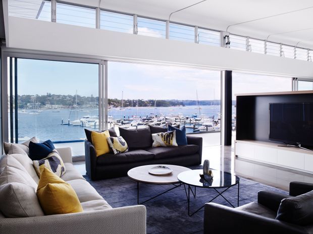 Bright, spacious contemporary living room with floor-to-ceiling glazing overlooking a marina and waterfront. The room features large sofas with navy and mustard cushions, round nesting coffee tables, a dark rug, and a wall-mounted television within white built-in cabinetry. High clerestory windows and a raised ceiling enhance the airy feel.