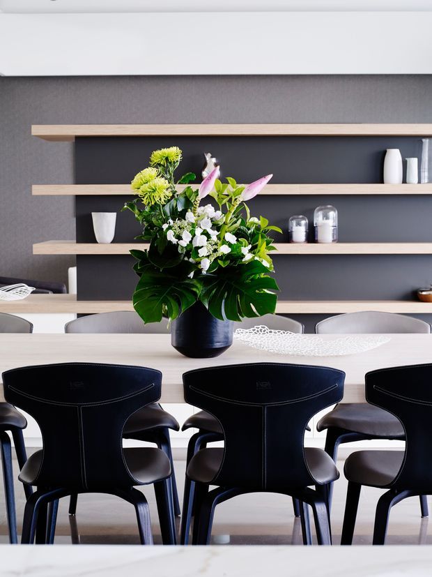 A refined dining area with a long pale timber dining table and dark upholstered dining chairs. A large floral arrangement in a dark vase forms the centrepiece, set against charcoal built-in shelving with pale timber shelves displaying ceramics and glassware. The palette is monochromatic with warm timber accents and contemporary styling.