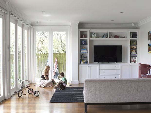 Bright family living room with polished timber flooring, extensive white-framed glazing and French doors opening to a garden, and a built-in white media and storage wall. A sofa faces the television, while children and toys occupy the light-filled space.