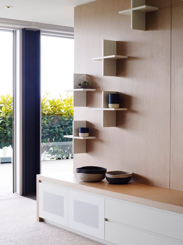 Bright contemporary interior featuring a built-in white storage console with a light timber benchtop, a full-height light timber feature wall with staggered open shelving, and a large glazed opening framed by a deep navy vertical panel. Decorative bowls and planters add restrained contrast.