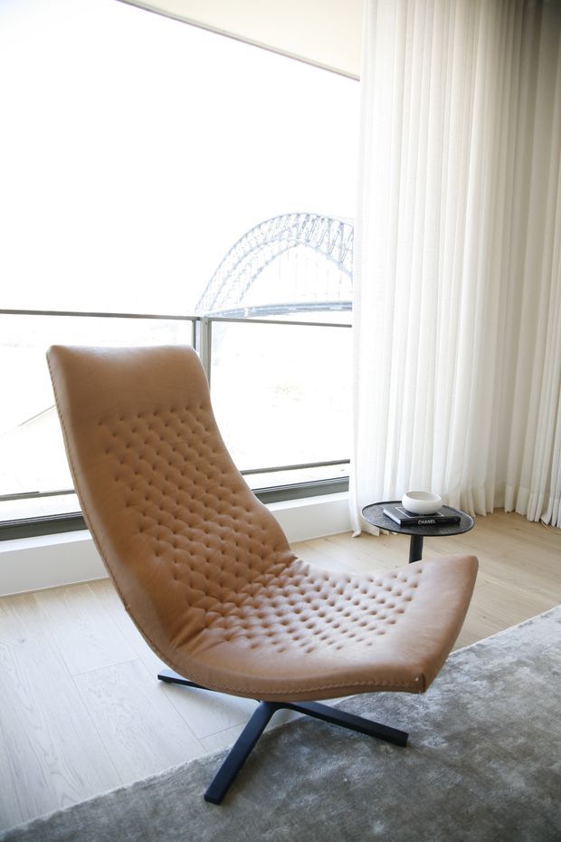 Bright contemporary living area featuring a tan tufted leather lounge chair on a dark cross-base, positioned beside floor-to-ceiling glazing with sheer white curtains. Pale timber flooring and a soft grey rug create a light, refined setting, with a small black side table holding books and a cup. A large arched bridge is visible through the window.
