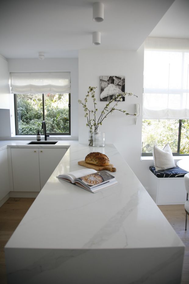 Bright, refined white kitchen centered on a large waterfall-edge stone island with subtle veining. The space includes white flat-panel cabinetry, a black undermount sink and tap, timber flooring, large windows with white Roman blinds, and a built-in window seat with patterned upholstery and cushions. Minimal decorative styling includes flowering branches, artwork, and an open cookbook on the island.