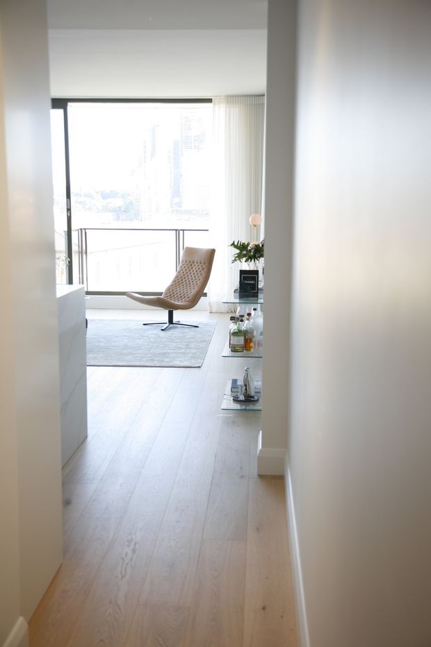 A bright, minimalist living room viewed from a hallway, featuring pale timber flooring, white walls and cabinetry, floor-to-ceiling glazing with vertical blinds, a large light rug, and a sculptural tan lounge chair. A small glass shelving unit displays bottles and decor, while an urban view is visible through the window.