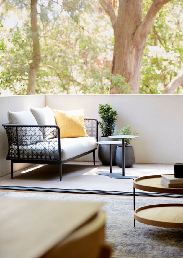 A bright, refined indoor living area opening onto a leafy terrace or courtyard. The space features a black-framed lounge chair with woven detailing and light cushions, a yellow accent cushion, round nesting side tables, potted greenery, and low circular timber coffee tables. Large glazing or an open threshold connects the interior to the sunlit outdoor seating area, with mature trees visible beyond.