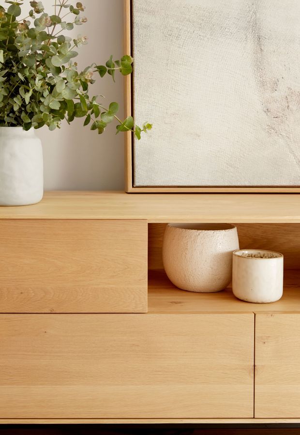 Close-up of a warm light-oak built-in console or sideboard with minimalist slab-front cabinetry and an open display niche. A white ceramic vase with leafy branches, neutral pottery, and a framed textured artwork add natural, understated styling.