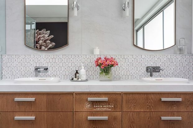 Bright contemporary bathroom vanity with a wide double-basin countertop, warm wood cabinetry, pale stone-look wall finish, and a patterned geometric tile splashback. Two wall-mounted mirrors, modern chrome sconces and tapware, and a floral arrangement complete the refined design.