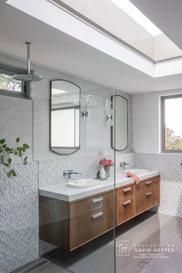 Bright contemporary bathroom with a double vanity in dark timber cabinetry, white benchtop and vessel basins, patterned geometric wall tiles, frameless glass shower screen, mirrored arch-top cabinets, and a skylight. A freestanding bathtub is partially visible at right.