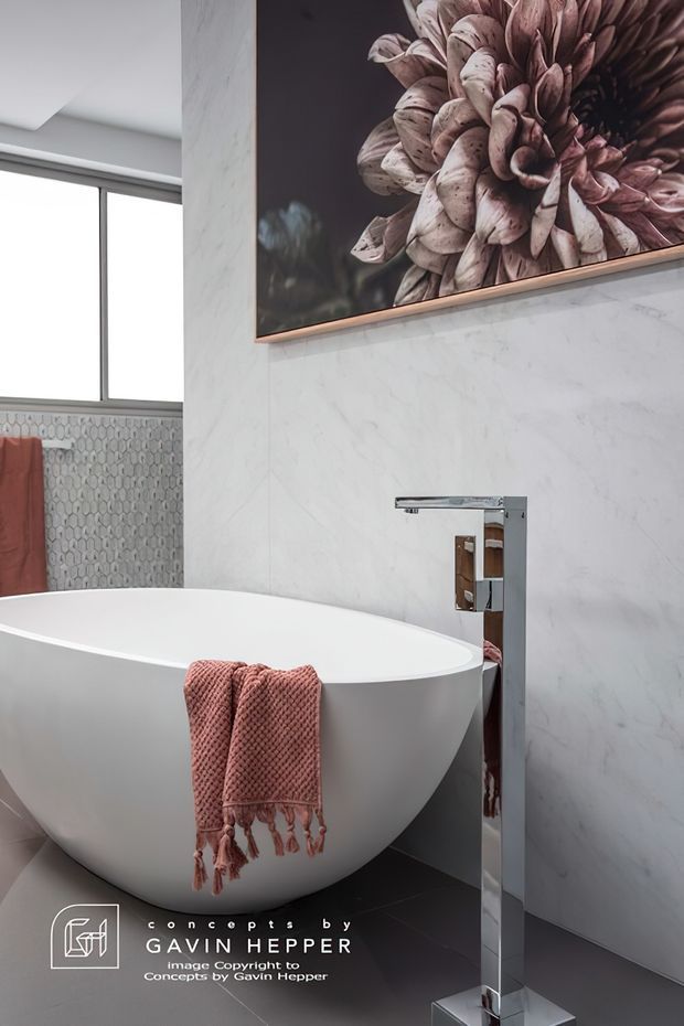 A refined contemporary bathroom featuring a freestanding white oval bathtub, a tall polished chrome floor-mounted tap, pale marble-look wall panels, dark grey floor tiles, and a large floral artwork above the bath. A patterned tiled wall, window, and blush-pink towels add texture and warmth.