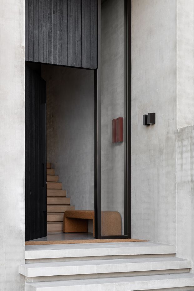 A minimalist contemporary entryway viewed from outside, framed by pale smooth concrete or plaster walls. Broad concrete steps lead to a tall black-framed glazed entrance with a dark vertically ribbed door panel. Inside, a timber staircase and curved brown built-in seating are visible, with understated red and black wall-mounted fixtures adding small accents.