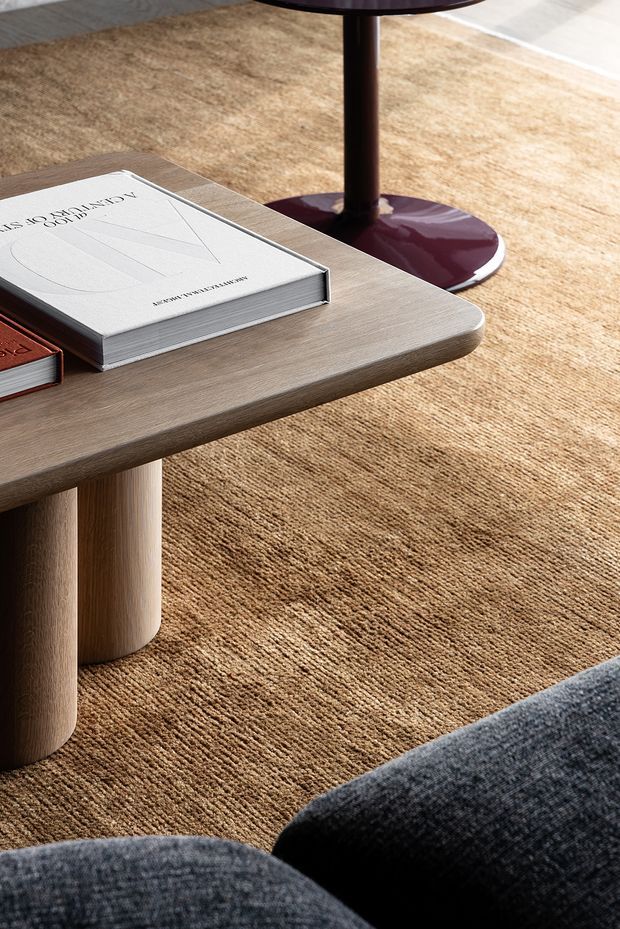 Close-up interior detail of a warm, contemporary living room, featuring a rounded light timber coffee table with books, a textured tan area rug, a dark upholstered sofa in the foreground, and a small burgundy pedestal side table.