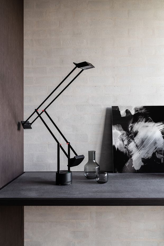Close-up interior vignette of a dark timber or wood-toned built-in shelving unit against a light textured brick wall. A black articulated desk lamp, glass vessels, and monochrome abstract artwork sit on the dark grey shelf surface, creating a restrained, graphic composition.