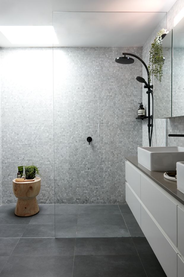 A contemporary bathroom with a walk-in glass shower, grey small-format circular mosaic wall tiles, large dark grey floor tiles, a white floating vanity with a grey benchtop, and a white vessel basin. Matte black shower fittings and tapware provide contrast, while a timber pedestal stool and hanging greenery add natural accents.