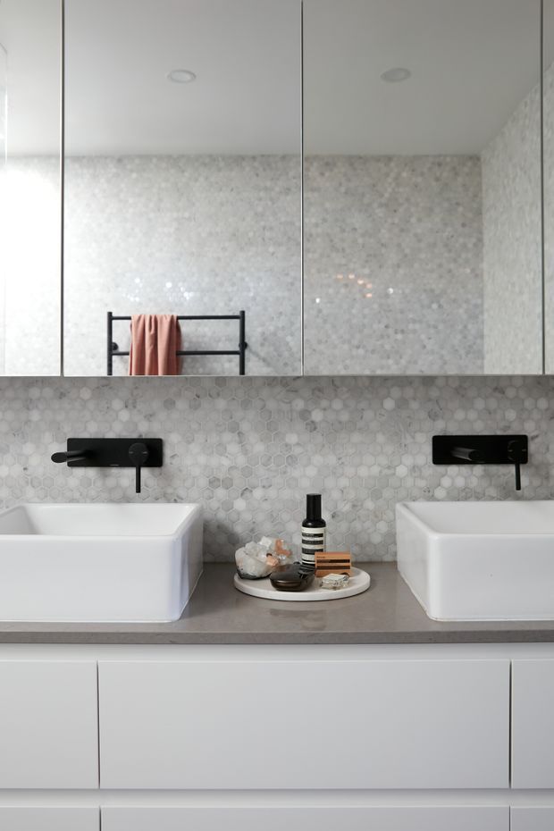 Contemporary double vanity bathroom featuring two white vessel basins, matte black wall-mounted faucets, a grey stone benchtop, white cabinetry, and a pale hexagonal mosaic tile splashback. Mirrored cabinets span the upper wall, reflecting the bathroom and a black towel rail with a blush towel.