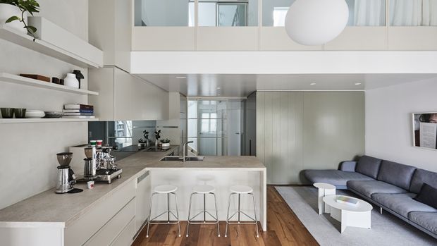 A bright, open-plan contemporary interior combining a compact kitchen with a central island and three stools, a lounge area with a sectional sofa, and a mezzanine-level glazed partition above. The space features pale cabinetry and walls, a stone-look benchtop, timber flooring, open shelving, and a large spherical pendant light.