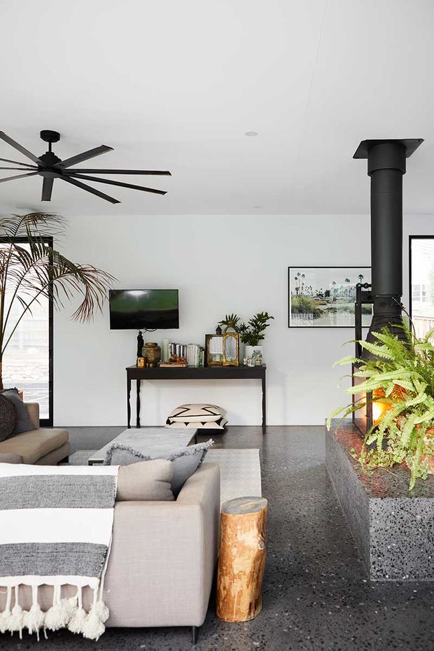 A bright contemporary living room with a large grey sectional sofa, layered neutral textiles, a black console table, wall-mounted television, indoor plants, framed artwork, and a prominent black freestanding wood-burning stove with flue. The room has a white ceiling and walls, polished dark terrazzo-style flooring, expansive glazing, and a large black ceiling fan. A raised built-in planter runs beside the stove and seating area.